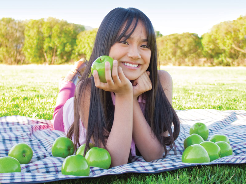 A young lady smiling outside on a blanket with apples.